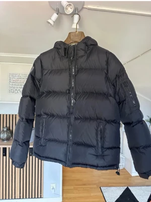 Svart dunjacka med huva från Peak Performance - Snygg svart dunjacka från Peak Performance med huva och dragkedja framtill. Jackan har två sidofickor med dragkedja och en ärmficka. Klassisk puffer-modell som håller dig varm under kalla dagar. Perfekt för vintern och enkel att matcha med olika outfits.