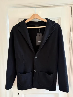 Svart Cardigan från SAND Copenhagen - Svart kofta från SAND Copenhagen med två stora fickor framtill och knäppning med två knappar. Koftan har en klassisk krage och rak passform. Materialet ser ut att vara mjukt och lite tjockare, vilket ger en skön och stilren känsla.