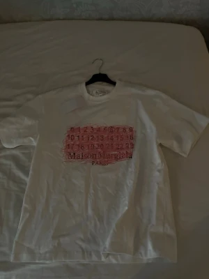 Vit Maison Margiela t-shirt - Vit t-shirt från Maison Margiela med tryck i rosa och svart framtill. Klassisk passform och rund hals. Perfekt nu till sommaren! Hör av er om ni vill ha fler bilde eller undrar något!