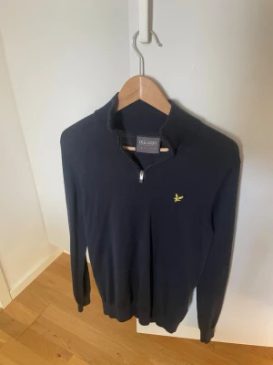 Svart halvzip-tröja från Lyle & Scott - Stilren svart långärmad tröja från Lyle & Scott med halv dragkedja och klassisk gul logga på bröstet. Tröjan har ribbade muddar och hög krage. Materialet känns mjukt och lätt, passar till många outfits. Använd 4/5 ggr