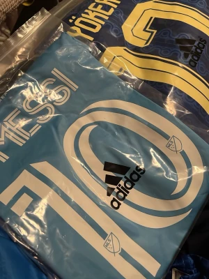 Ljusblå Messi fotbollströja Adidas - Säljer en ljusblå fotbollströja från Adidas med Messi och nummer 10 tryckt på ryggen. Tröjan har vita detaljer och MLS-logga, perfekt för dig som älskar fotboll och vill bära din idol på ryggen.