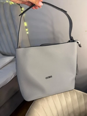 Ljusgrå handväska från Guess - Snygg ljusgrå handväska från Guess i stilren design. Väskan har en bred axelrem och är tillverkad i strukturerat syntetmaterial. Guess-loggan i silver framtill ger en clean look. Perfekt storlek för vardagens essentials. Köpt från Guess och aldrig använt helt ny med prislapp
