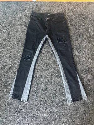Svarta jeans med slitningar BoohooMAN - Unika svarta jeans från BoohooMAN med ljusgrå paneler längs benen och slitna detaljer på låren. Modellen har raka ben med fransade benslut och klassisk femficksdesign. Perfekta för dig som vill sticka ut med en edgy streetstyle.