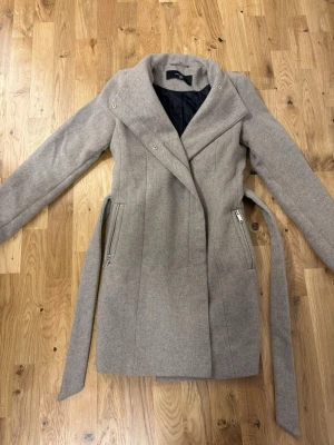 Beige kappa  - Stilren lång beige kappa från Zara med bred krage, snedställd dragkedja och går att knyta i midjan. I väldigt bra skick och använd fåtal gånger. Kappan har två fickor med dragkedja och ett svart innerfoder. 