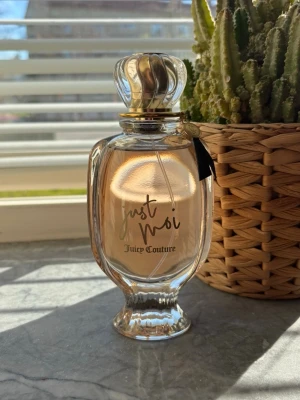 Just Moi parfym från Juicy Couture - Elegant parfymflaska i genomskinligt glas med guldiga detaljer och texten 'Just Moi'. Doften kommer från Juicy Couture och flaskan har en lyxig, klassisk form med dekorativ kork. 100ml, ny