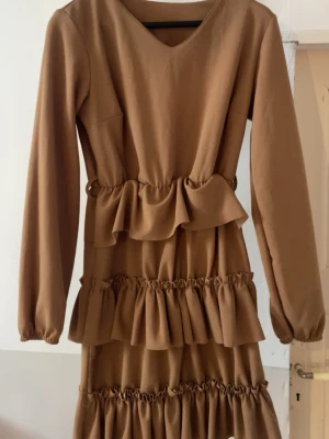 Brun volangklänning med lång ärm - Snygg brun klänning med flera volangdetaljer och lång ärm. Klänningen har v-ringning och markerad midja med volanger som ger en trendig look. Perfekt för dig som gillar en feminin och modern stil.