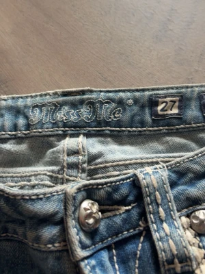 Bootcut Miss Me ljusblå jeans med detaljer - Snygga ljusblå bootcut jeans från Miss Me med dekorativa kontrastsömmar och unika fickdetaljer med knappar och broderier. Jeansen har klassisk femficksdesign och coola detaljer på bakfickorna. Perfekta för dig som gillar statement-jeans.