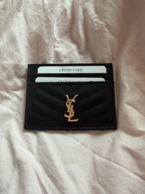 Svart kortfodral från Yves Saint Laurent - Snyggt svart kortfodral från Saint Laurent i quiltat läder med guldfärgad YSL-logga framtill. Fodralet har flera fack för kort och en stilren, minimalistisk design som passar perfekt i fickan eller väskan. Ej äkta!