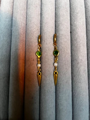 Guldiga örhängen med grön sten och pärla - Snygga guldiga örhängen med en grön rund sten, liten vit pärla och spetsig hängande detalj. Perfekt accessoar för att lyfta din stil med en cool och elegant touch. Materialet ser ut att vara metall med färgade stenar och pärldetalj.