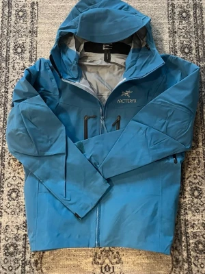 Arc'teryx jacka - Snygg blå rengjacka/vindjacka från Arc'teryx med huva och dragkedja framtill. Jackan har två bröstfickor med dragkedja och diskret logga på bröstet. Perfekt för blåsiga dagar och outdoor-aktiviteter. Materialet är lätt och vattenavvisande. Pris kan diskuteras vid snabb affär.
