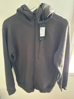 Mörkgrå hoodie från Sail Racing - Mörkgrå hoodie från Sail Racing med hel dragkedja och huva med dragsko. Materialet är mjukt och bekvämt, perfekt för chill dagar. Bröstficka med dragkedja och diskret logotyp på bröstet. Långa ärmar och ribbade muddar för extra komfort. Säljs till ett otroligt bra pris helt ny 499kr 