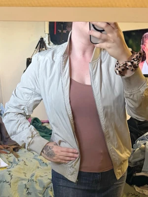Beige bomberjacka med ribbade muddar - Stilren beige bomberjacka med dragkedja framtill och ribbade muddar vid ärmslut och midja. Jackan har klassisk bombermodell med rundad krage och lätt glansigt material. Perfekt för en trendig och avslappnad look.