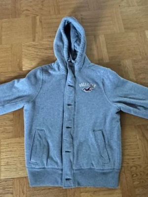 Grå hoodie från Hollister med knappar - Snygg grå hoodie från Hollister med quiltat foder och knäppning framtill. Hoodien har två fickor, broderad logga på bröstet och en mysig huva. Perfekt för dig som gillar en avslappnad och stilren look. Perfekt nu till sena sommarkvällar kan även användas till vår jacka!☀️ användt några gånger mest legat. Vintage Hollister hoddie säljs inte längre. Denna moddel samt färg är väldigt unik. Pris går att diskutera! Mått finns vid intresse! Storlek L
