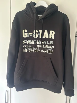 Svart hoodie från G-Star RAW - Svart hoodie från G-Star RAW med stor vit logga och tryck på bröstet. I väldigt fint skick. Storlek M. Pris 149kr
