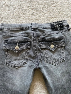 Gråa True Religion jeans med fickdetaljer - Snygga grå jeans från True Religion med ikoniska bakfickor och kontrastsömmar. Jeansen har lock på bakfickorna med silverfärgade knappar och en svart patch med logga i midjan.
