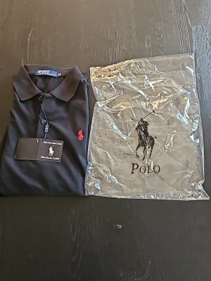 Svart pikétröja från Polo Ralph Lauren - Klassisk svart pikétröja från Polo Ralph Lauren med röd broderad logga på bröstet. Tröjan har krage, korta ärmar och knäppning framtill. Perfekt för en stilren och avslappnad look.