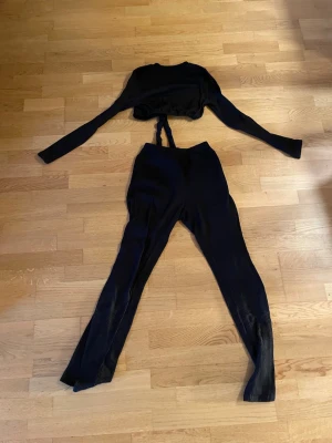 Svart ribbat set med långärmad topp - Snyggt svart tvådelat set med långärmad crop top och höga ribbade byxor. Setet har en tight passform och är gjort i stretchigt material som sitter skönt på kroppen. Perfekt för en trendig och bekväm look.