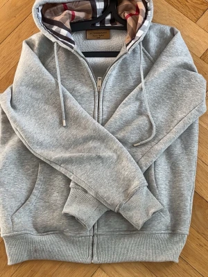 Grå zip-hoodie från Burberry - Snygg grå hoodie från Burberry med dragkedja och klassiskt rutigt foder i huvan. Mjukt material, ribbade muddar och fickor framtill. Perfekt för en chill och stilren look.