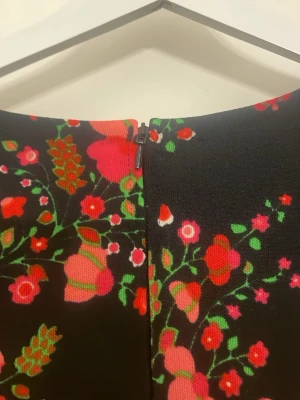 Svart långklänning med blommigt mönster - Långklänning med svart bas och färgglatt blommigt mönster i rosa, rött och grönt. Klänningen har v-ringning, korta ärmar och en flowy passform. Perfekt för dig som vill sticka ut med en romantisk och trendig look.