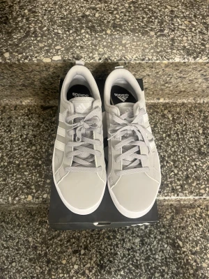 Gråa Adidas sneakers med vita ränder - Snygga gråa sneakers från Adidas med klassiska vita ränder på sidorna och vit sula. Skorna har en låg profil, snörning och är tillverkade i syntetmaterial med textilfoder. Perfekta för en stilren och sportig look.