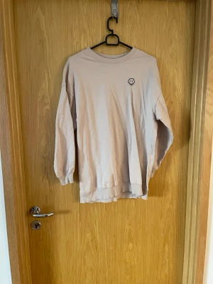 Beige sweatshirt med smiley från H&M - Säljer en beige sweatshirt från H&M i storlek XS. Tröjan har en enkel design med en liten broderad smiley på bröstet. Tillverkad i mjukt bomullsmaterial och har ribbade muddar vid ärmslut och hals. Perfekt för en chill och avslappnad stil.