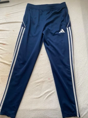 Blå Adidas träningsbyxor - Mörkblå träningsbyxor från Adidas med vita klassiska ränder längs benen och Adidas-logga på låret. Byxorna har resår i midjan och smal passform, perfekta för sport eller chill. Tillverkade i ett lätt och stretchigt material.