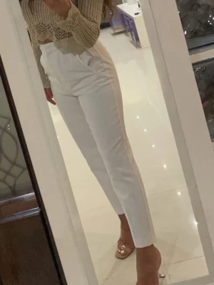 Vita höga kostymbyxor - Säljer ett par vita kostymbyxor med hög midja och avsmalnande ben. Byxorna har en clean look utan synliga fickor eller detaljer och är perfekta för en stilren outfit. Materialet ser ut att vara mjukt och följsamt, troligen polyester eller bomullsblandning.