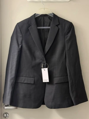 Mörkgrå klassisk kavaj - Stilren mörkgrå kavaj med enkelknäppning och två fickor framtill. Klassisk krage och figurnära passform. Perfekt för en snygg och tidlös look. 