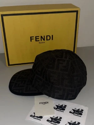 Svart keps med Fendi-logga - Snygg svart keps från Fendi med det klassiska FF-monogrammet över hela kepsen. Kepsen har böjd skärm och är tillverkad i ett tygmaterial som känns lyxigt. Perfekt accessoar för att lyfta din streetwear-look.