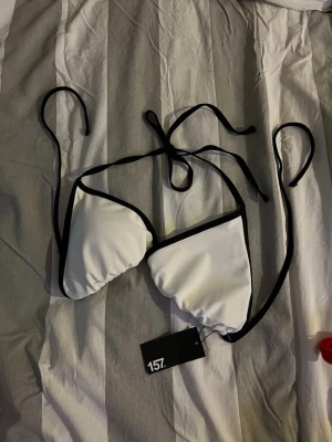 Vit och svart bikiniöverdel 157 - Snygg trekantsbikini från 157 i vit med svarta kanter och knytband i nacken och ryggen. Enkel och stilren design som passar perfekt till sommaren. Materialet känns mjukt och bekvämt mot huden.