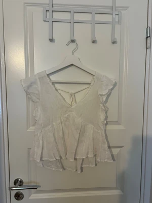 Vit blus med volang och knyt - Söt vit blus med v-ringning, volangkant och knyt i ryggen. Blusen har korta ärmar och är tillverkad i ett lätt, luftigt tyg som passar perfekt till sommaren. Den har en avslappnad passform och fina detaljer längs kanterna.