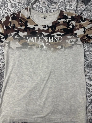 Valentino camo t-shirt i bomull - Snygg t-shirt från Valentino Garavani med camouflagemönster i brunt, svart och beige upptill och ljusgrå nederdel. Stor logga framtill. Tillverkad i mjuk bomull och har klassisk rund hals och korta ärmar. Perfekt för en trendig och avslappnad stil.