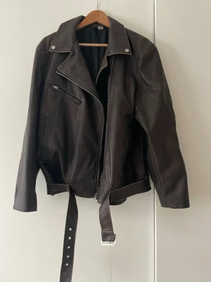 Brun biker skinnjacka med bälte - Snygg brun skinnjacka i klassisk bikerstil med bred krage, silverfärgade knappar och dragkedja. Jackan har ett bälte i midjan, snedställd bröstficka med dragkedja och två sidofickor. Perfekt för dig som vill ha en tidlös och cool look oversize modell, så något stor i storleken.
