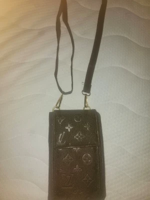 Svart plånbok med axelrem - Snygg brun plånbok med klassiskt monogrammönster i präglat läder. Plånboken har en praktisk axelrem och ett lock med tryckknapp. Perfekt för att bära det viktigaste och samtidigt hålla stilen.
