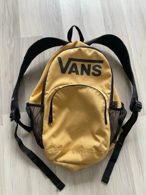 Gul ryggsäck från Vans - Snygg gul ryggsäck från Vans med svart logga framtill. Ryggsäcken har ett stort huvudfack, en mindre ficka framtill med dragkedja och två sidofickor i mesh. Justerbara svarta axelband och praktisk ögla upptill. Perfekt för skolan eller utflykter.