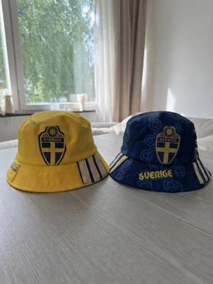 Sverige bucket hats gul och blå - Två bucket hats med Sverige Fotbolls landslagsmärke. En hatt är gul med blå och vita ränder, den andra är blå med gula detaljer och texten 'Sverige'. Perfekta för att visa ditt stöd under matchdagar eller festivaler.