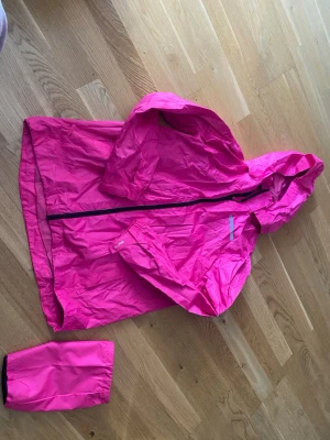 Rosa regnjacka med huva - Snygg rosa regnjacka med huva och svart dragkedja. Jackan är lätt och smidig, tillverkad i vattenavvisande material och har resår vid ärmsluten. Praktisk att packa ner i medföljande påse. Perfekt för regniga dagar.