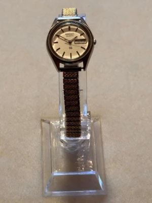 Seiko herrklocka i silver och guld - Klassisk Seiko Quartz herrklocka med rund urtavla i silver och dag/datumvisning. Armbandet är i metall med inslag av både silver och guldtoner, vilket ger en tidlös och stilren look. Perfekt accessoar för dig som gillar vintagevibbar och enkel design.