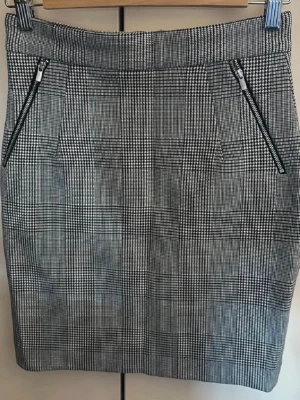 Rutig pennkjol med dragkedjor från H&M - Snygg rutig kortkjol i svart och vitt med två dragkedjor framtill. Kjolen har en klassisk pennkjolsform och hög midja, perfekt för en stilren look. Materialet känns som en blandning av polyester och bomull. OBS: FRAKTAR INOM 24H!🩷