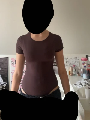 Brun basic body från H&M - Säljer en enkel och stilren brun body från H&M med korta ärmar och rund halsringning. Tillverkad i ett mjukt och stretchigt material som sitter tajt mot kroppen. Perfekt att styla med jeans eller kjol för en clean look.