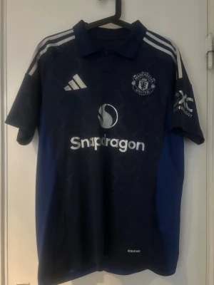 Blå Manchester United matchtröja Adidas - Snygg blå Manchester United matchtröja från Adidas med vita detaljer och loggor. Tröjan har korta ärmar, krage och tryckta sponsorer som Snapdragon. Tillverkad i lätt och ventilerande material med Aeroready-teknologi, perfekt för träning eller match.