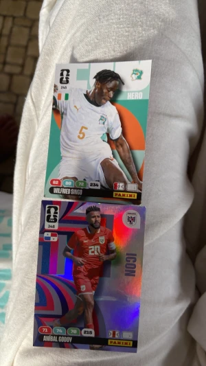 Panini fotbollssamlarbilder - Två Panini fotbollssamlarbilder: Wilfried Singo i vit landslagströja från Elfenbenskusten och Aníbal Godoy i röd landslagströja från Panama. Båda korten har färgstark design och visar spelarnas statistik. Perfekt för samlare eller fotbollsfans.
