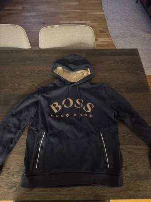 Hugo boss hoodie - Snygg svart hoodie från Hugo Boss med stor logga i guld på bröstet. Hoodien har dragsko i huvan och kontrastfärgad insida i beige. Praktiska fickor med dragkedja på sidorna. Perfekt för en chill och stilren look. Passar M också