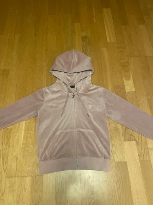 Rosa velourhoodie från Juicy Couture - Mjuk och lyxig rosa velourhoodie från Juicy Couture med dragkedja, huva med dragsko och JC-broderi på bröstet. Perfekt för en chill och trendig look. Stora fickor framtill och ribbade muddar ger extra komfort.