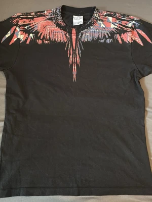 Svart t-shirt från Marcelo Burlon - Svart t-shirt från Marcelo Burlon med ikoniskt färgstarkt vingmönster i rött, blått och vitt över axlar och halsringning. Klassisk passform och rund hals. Perfekt statement-plagg för dig som vill sticka ut.