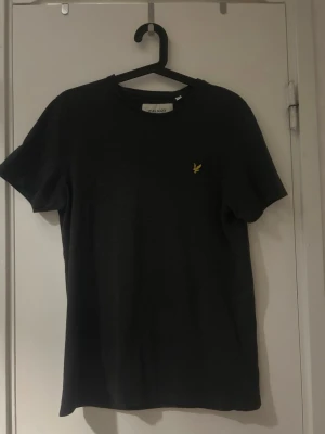 Mörkgrå  t-shirt från Lyle & Scott - Klassisk svart t-shirt från Lyle & Scott med gul logga på bröstet. T-shirten har rund halsringning och korta ärmar. Perfekt basplagg med enkel och stilren design.