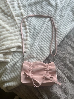 Ljusrosa axelremsväska med rosetter - Söt ljusrosa handväska i sammetsliknande material med justerbar axelrem. Väskan har rynkade detaljer och två dekorativa rosetter framtill. Magnetlås och innerfack med dragkedja för smidig förvaring. Perfekt accessoar för en trendig look.
