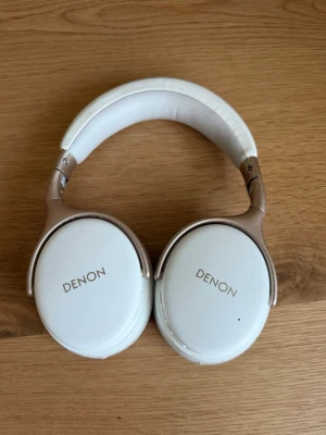 Vita over-ear hörlurar från Denon - Snygga vita over-ear hörlurar från Denon med mjuka, ovala öronkuddar och justerbart huvudband. Hörlurarna har en stilren design med gulddetaljer och vadderad bygel för extra komfort. Perfekta för musik, gaming eller poddar.