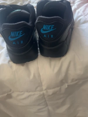 Nike Air Max 90 svart/blå sneakers - Säljer ett par svarta Nike Air Max 90 sneakers med blå detaljer och klassisk Air-dämpning i sulan. Skorna har en låg silhuett, ovandel i en mix av läder och mesh samt tydliga Nike-loggor på häl och sida. Perfekta för dig som gillar streetwear och sportig stil.