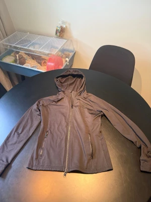 Svart ridtröja från Fairfield - Snygg svart ridtröja från Fairfield med huva och dragkedja framtill. Jackan har två fickor med dragkedja och är tillverkad i ett lätt, syntetiskt material. Perfekt för blåsiga dagar och enkel att matcha med olika outfits.